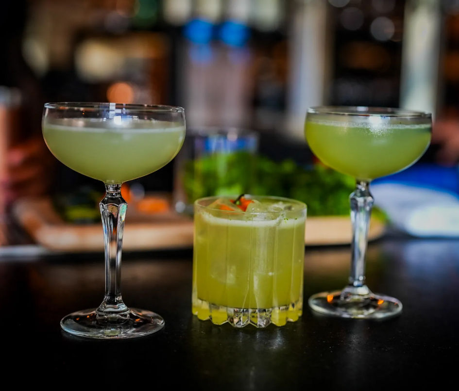 Three vibrant green cocktails on a bar: two in coupe glasses and one in a short rocks glass with ice and a small red garnish, set against a blurred bar background.