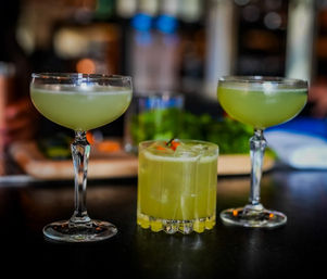 Three vibrant green cocktails on a bar: two in coupe glasses and one in a short rocks glass with ice and a small red garnish, set against a blurred bar background.