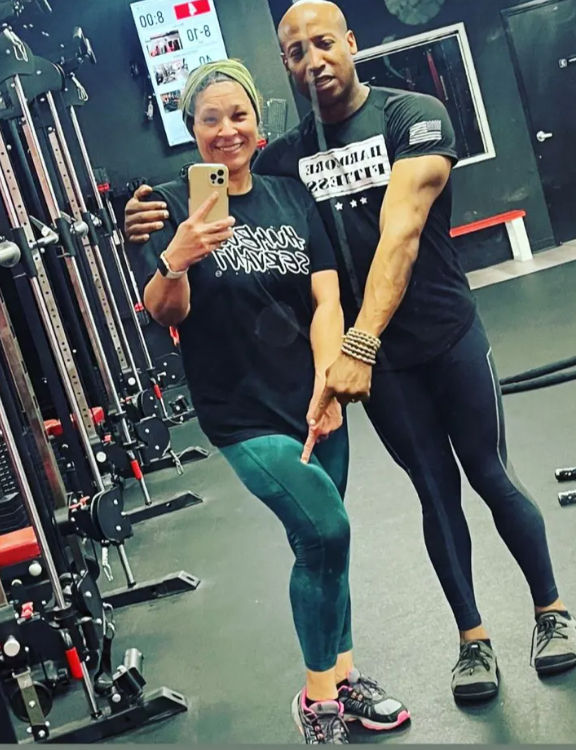 Mirror selfie in a gym: two people in athletic wear — one smiling and holding a phone, the other pointing at a leg — standing by cable machines and benches in a fitness studio.