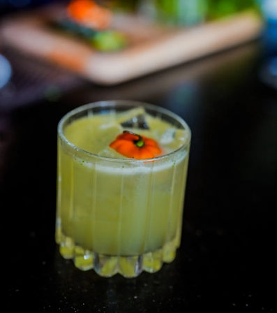 Chilled green craft cocktail in a short ribbed glass with ice and a bright orange edible flower garnish, sitting on a dark bar surface.