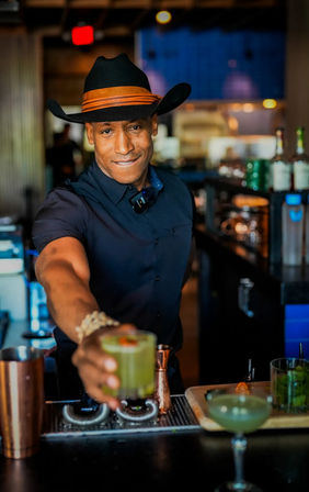 Smiling bartender in a black cowboy hat and navy shirt reaches forward to hand a bright green craft cocktail garnished with a small red pepper at a modern urban cocktail bar.