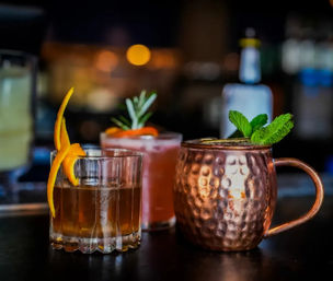 Three craft cocktails on a dim bar counter: an old fashioned with an orange twist, a pink rosemary cocktail, and a copper Moscow mule garnished with fresh mint.