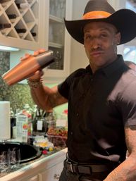 Person in a cowboy hat and black shirt shaking a copper cocktail shaker in a home kitchen bar, with a wine rack, fruit and glassware on the countertop.