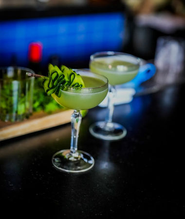 Two vibrant green cucumber cocktails in coupe glasses on a dark bar counter, each topped with a curled cucumber ribbon garnish — refreshing craft cocktail.