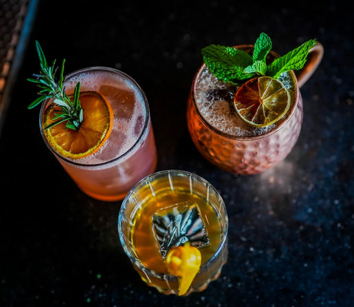 Top-down view of three colorful craft cocktails on a dark bar surface — pink citrus drink garnished with rosemary and dried orange, a copper mug with mint and lime, and a lowball glass with a large ice cube and citrus twist.