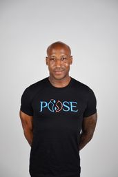 Studio headshot of a bald man wearing a black t-shirt with a blue-orange graphic logo, tattooed forearms and a friendly smile against a light gray background