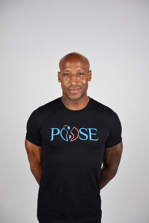 Studio headshot of a bald man wearing a black t-shirt with a blue-orange graphic logo, tattooed forearms and a friendly smile against a light gray background