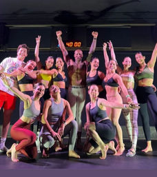 Energetic group fitness class posing on a studio stage, instructor at center with arms raised and a 40:00 timer overhead, smiling participants in workout gear.