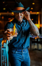 Person in a black cowboy hat tipping the brim and offering an amber cocktail with an orange peel garnish in a cozy dimly lit bar lounge.