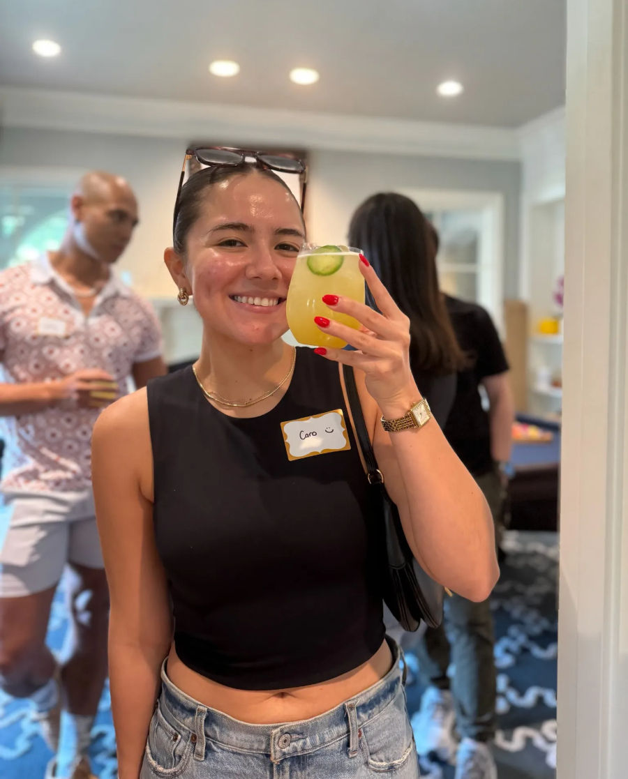 Smiling woman in a black crop top and denim jeans holding a yellow cucumber‑garnished cocktail at a casual indoor house party with guests in the background.