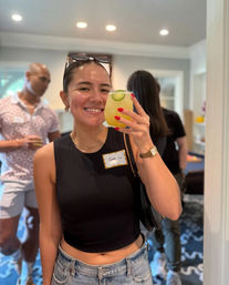 Smiling woman in a black crop top and denim jeans holding a yellow cucumber‑garnished cocktail at a casual indoor house party with guests in the background.