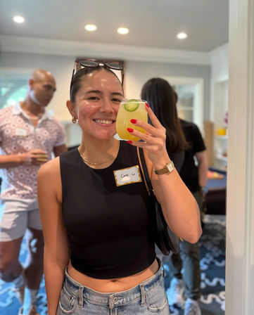 Smiling woman in a black crop top and denim jeans holding a yellow cucumber‑garnished cocktail at a casual indoor house party with guests in the background.