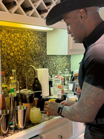 Home bartender in a black cowboy hat crafting a citrus cocktail at a granite kitchen counter with metal shakers, strainer, juice carton and a glass of pale yellow drink.