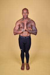 Shirtless athletic man with chest and arm tattoos and beaded bracelets stands barefoot on a yellow studio backdrop in a namaste prayer pose wearing black compression leggings.