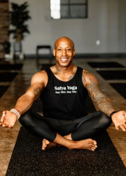Smiling fit man with tattoos seated cross‑legged on a yoga mat in an indoor studio, wearing black leggings and a tank top with a yoga slogan, palms open in meditation.