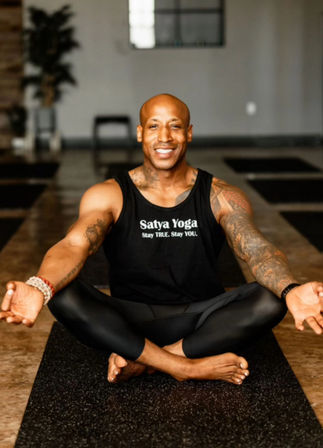 Smiling fit man with tattoos seated cross‑legged on a yoga mat in an indoor studio, wearing black leggings and a tank top with a yoga slogan, palms open in meditation.