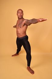 Bare-chested, tattooed man in black compression leggings holding a Warrior II yoga pose on a warm yellow studio backdrop.