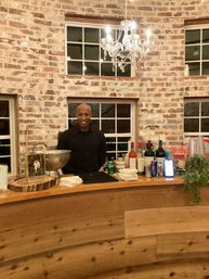 Smiling bartender standing behind a curved wooden bar stocked with wine, beer and soda, in a rustic indoor event space with exposed brick walls, window panes and a sparkling crystal chandelier overhead.