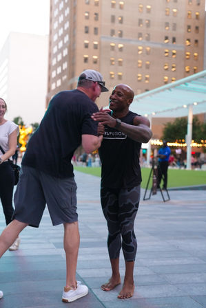 Two men share a friendly handshake and hug in a downtown city plaza at dusk, one barefoot in athletic leggings and the other in shorts and sneakers, with tall buildings and a covered lawn area and string lights in the background.