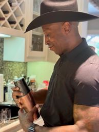 Person in a black cowboy hat and black shirt smiling while shaking a copper cocktail shaker in a bright home kitchen with wine rack and glassware on the counter.
