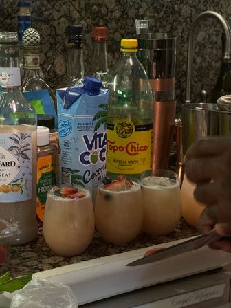 Three creamy pink cocktails garnished with sliced strawberries and blueberries on a granite kitchen counter, surrounded by a coconut water carton, sparkling mineral water, syrups and cocktail tools while a hand slices fruit.