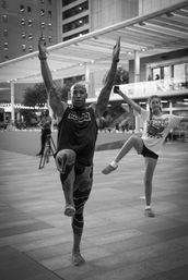 Athletic instructor leading an outdoor fitness class in a modern downtown plaza, balancing barefoot on one leg with arms raised while a participant mirrors the move under a glass canopy.
