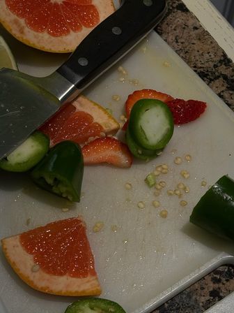 Kitchen cutting board with zesty grapefruit wedges, sliced strawberries and jalapeño rounds, a chef's knife and scattered seeds on a countertop