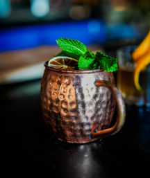 Hammered copper mug Moscow Mule cocktail topped with fresh mint sprig and a dried lime wheel, sitting on a dark bar counter with vibrant blue bokeh lights