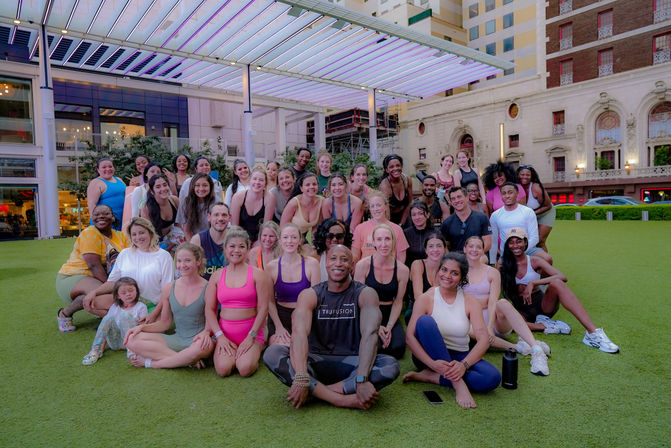 Large group of smiling people in colorful workout clothes posing on a green urban lawn under a modern lit pergola, instructor seated front center with city buildings and a theater-style facade in the background — outdoor group fitness class in a downtown plaza.