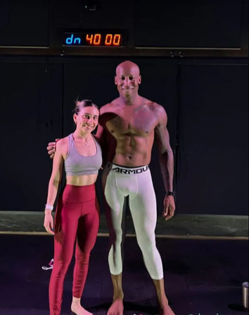 Two fit people posing barefoot in a dim fitness studio under a digital timer reading 40:00 — woman in a gray sports bra and red leggings and a shirtless man in white compression tights with a dark waistband, smiling after a workout.