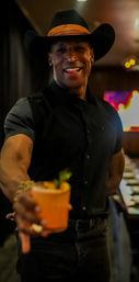 Smiling man in a black cowboy hat and shirt offers a bright orange craft cocktail toward the camera in a cozy, dimly lit cocktail bar.