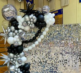 Black, silver and white balloon garland with disco-ball and star accents framing a mirrored sequin photo wall in a school gymnasium event