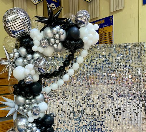 Black, silver and white balloon garland with disco-ball and star accents framing a mirrored sequin photo wall in a school gymnasium event