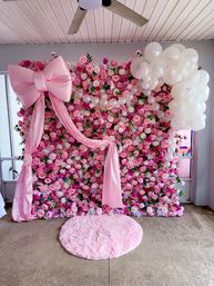 Indoor pink rose wall photo backdrop with large satin bow and draped fabric, cascading white balloon cluster, and round fluffy pink rug