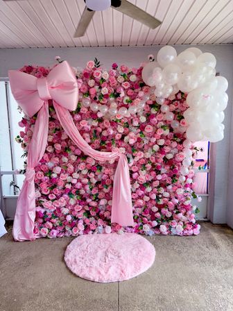 Indoor pink rose wall photo backdrop with large satin bow and draped fabric, cascading white balloon cluster, and round fluffy pink rug