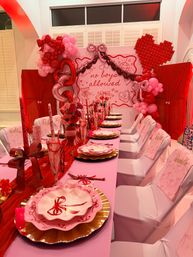 Indoor pink-and-red girls-only party table: layered bow-patterned pink plates on gold chargers, red ribbon centerpieces, pink chair covers, heart and balloon garlands and a backdrop reading 'no boys allowed'.