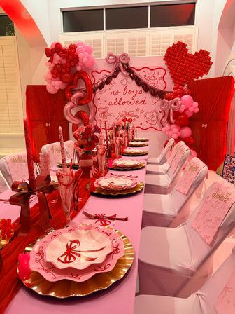 Indoor pink-and-red girls-only party table: layered bow-patterned pink plates on gold chargers, red ribbon centerpieces, pink chair covers, heart and balloon garlands and a backdrop reading 'no boys allowed'.