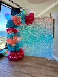 Vibrant balloon arch in teal, coral and magenta with iridescent spheres framing a shimmering turquoise sequin backdrop by a floor-to-ceiling window overlooking blue coastal water — party photo booth on wood floor.