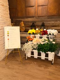 Indoor flower station with vibrant sunflowers, red and orange roses, white daisies and greenery in white pots on a wheeled cart against a rustic wood and metal wall, next to a gold easel sign
