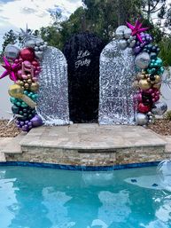 Poolside backyard party photo backdrop with a black sequin arch reading "Let's Party" between silver sequin panels, framed by colorful metallic balloon garlands, disco balls and star-shaped balloons.