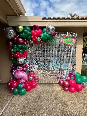 Tropical poolside patio party backdrop with mirrored sequin wall, vibrant pink and green balloon arch, metallic disco balls and a flamingo foil balloon.