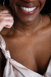 Close-up of a person with a sparkling smile wearing a delicate gold chain necklace and knotted silk camisole, focused on the collarbone and neckline.