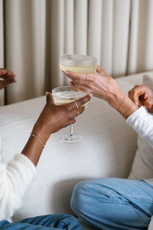 Two people clinking champagne coupe glasses over a cream sofa in a cozy living room, casual jeans and delicate gold bracelets visible