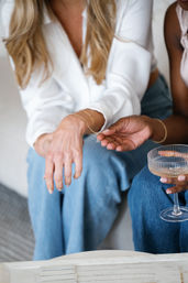 Close-up of two friends on a couch trying on a delicate gold bracelet, one hand holding the chain while the other wears a white blouse and jeans, with a sparkling coupe and jewelry tray nearby.