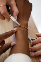 Close-up of hands using needle-nose pliers to adjust a delicate gold chain bracelet on a wrist, jewelry repair and bracelet sizing on a wooden tabletop.