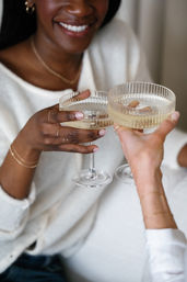 Close-up of two friends clinking ribbed champagne coupe glasses filled with sparkling wine, smiling faces and hands adorned with delicate gold jewelry in a cozy neutral setting.