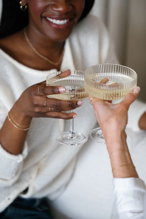 Close-up of two friends clinking ribbed champagne coupe glasses filled with sparkling wine, smiling faces and hands adorned with delicate gold jewelry in a cozy neutral setting.
