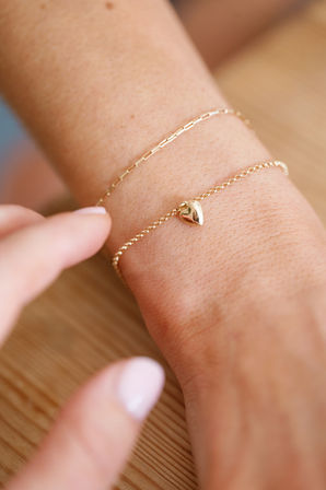 Close-up jewelry shot: dainty gold heart charm bracelet stacked with a thin gold chain on a wrist over a light wood surface