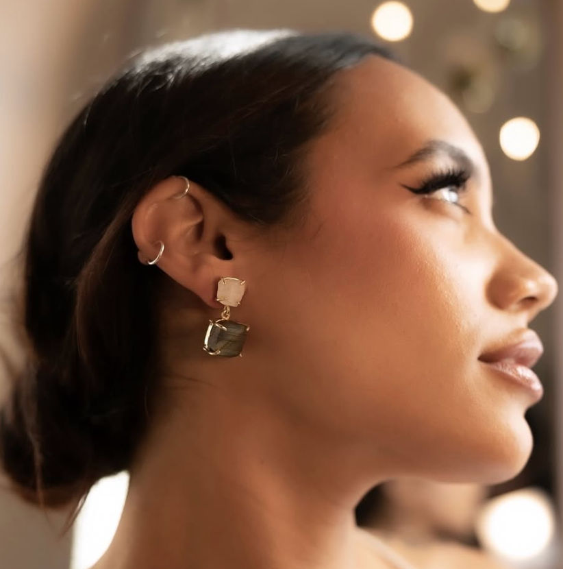 Close-up profile portrait of a woman wearing gold gemstone drop earrings and ear cuffs, warm indoor lighting with soft bokeh lights