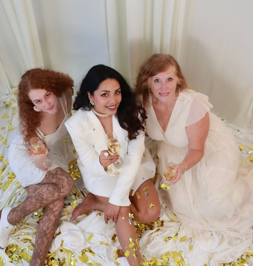 Three women in white dresses sitting on draped white fabric with gold confetti, smiling and toasting with champagne glasses in an indoor celebratory photoshoot.